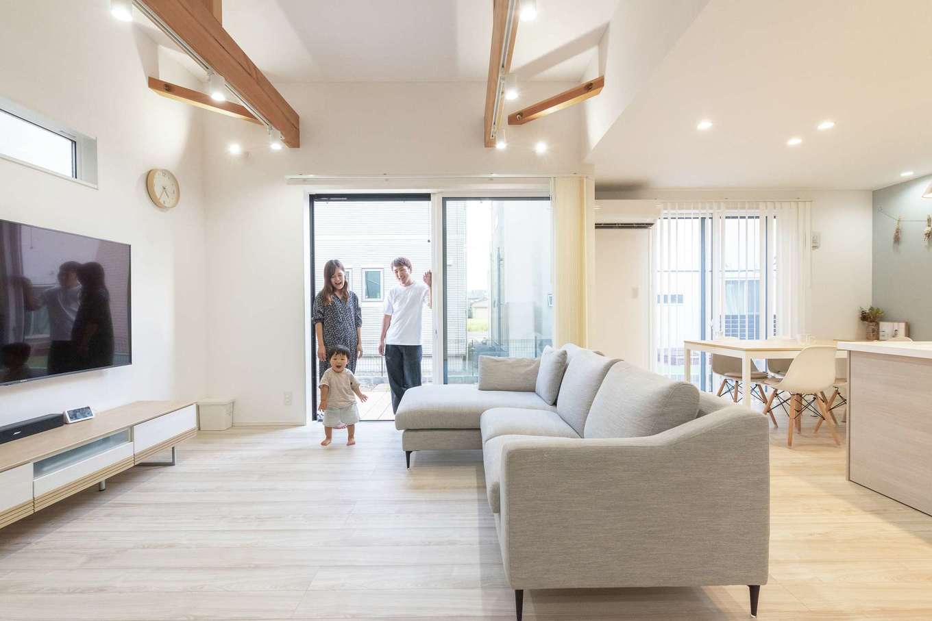 HAPPY HOUSE（山田工務店）【デザイン住宅、子育て、間取り】開放的なリビングは、優先順位の最上位だった。吹き抜けにすることも考えたが、2階の間取りと費用面から、天井を高くするという同社からの提案を採用した。床や壁に加え、インテリアやブラインドも、広く見せる明るい色をベースにコーディネート。窓の先はデッキと庭がつながり、さらなる開放感がもたらされた。当初は窓際に階段が計画されたが、「必要以上にキッチン前を横切るのはちょっと」という奥さまの意見で、LDKの入口付近に配置。右手奥のブルーのクロスの下には宿題にピッタリのデスクが設けられている