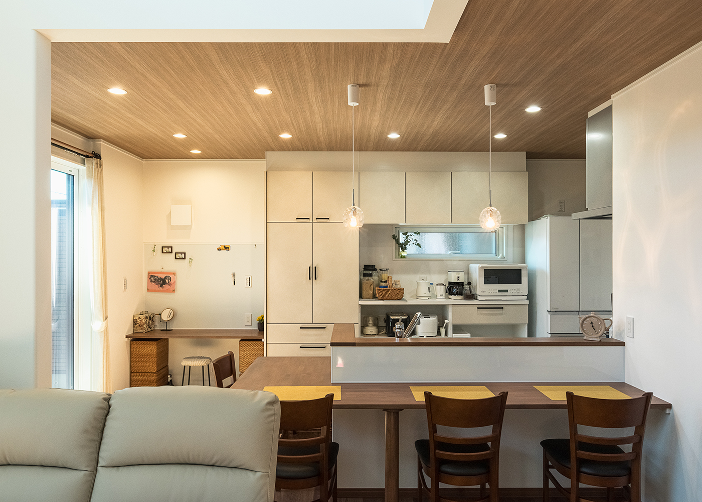イシカワ【収納力、省エネ、ペット】ダイニングテーブル一体型の対面キッチン。木目調の下がり天井がニュアンスを与える。キッチンの背面は、食器棚、調理家電、冷蔵庫まですべて白で統一した。壁のRの出隅は標準仕様で、安全性を保ちつつ優しさを表現している