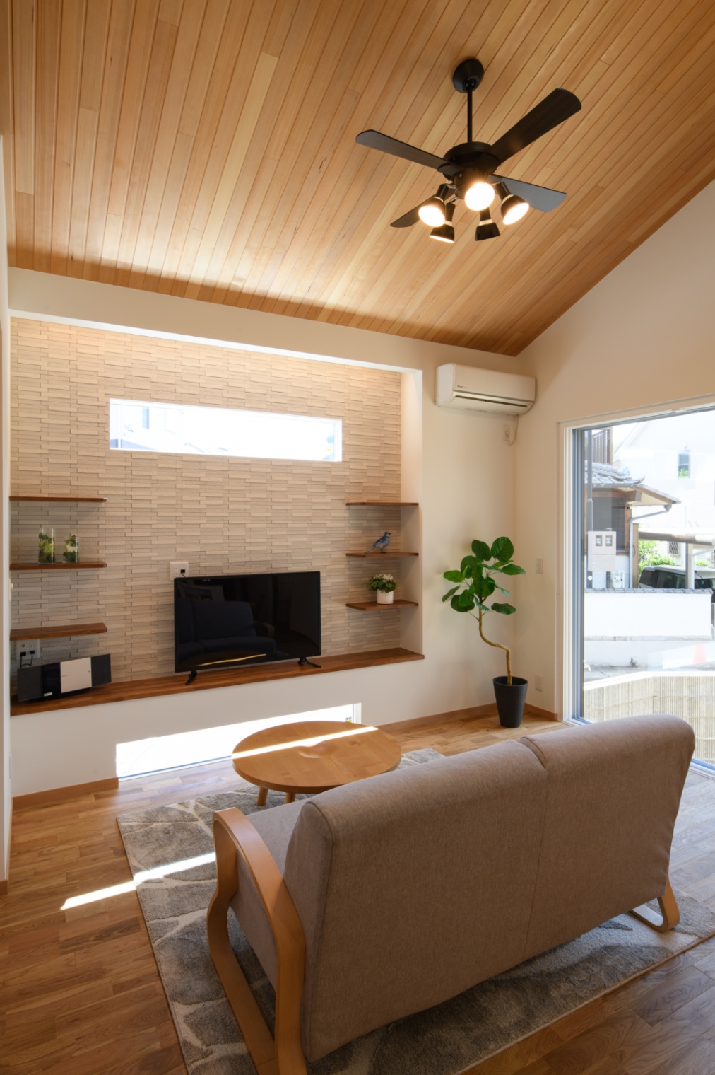 Sus∞Eco Home（サスエコホーム）｜カジュアルなリビングスペースで、木目天井と自然光が心地よい。シンプルな家具が配置され、明るい雰囲気の中でリラックスできる空間。