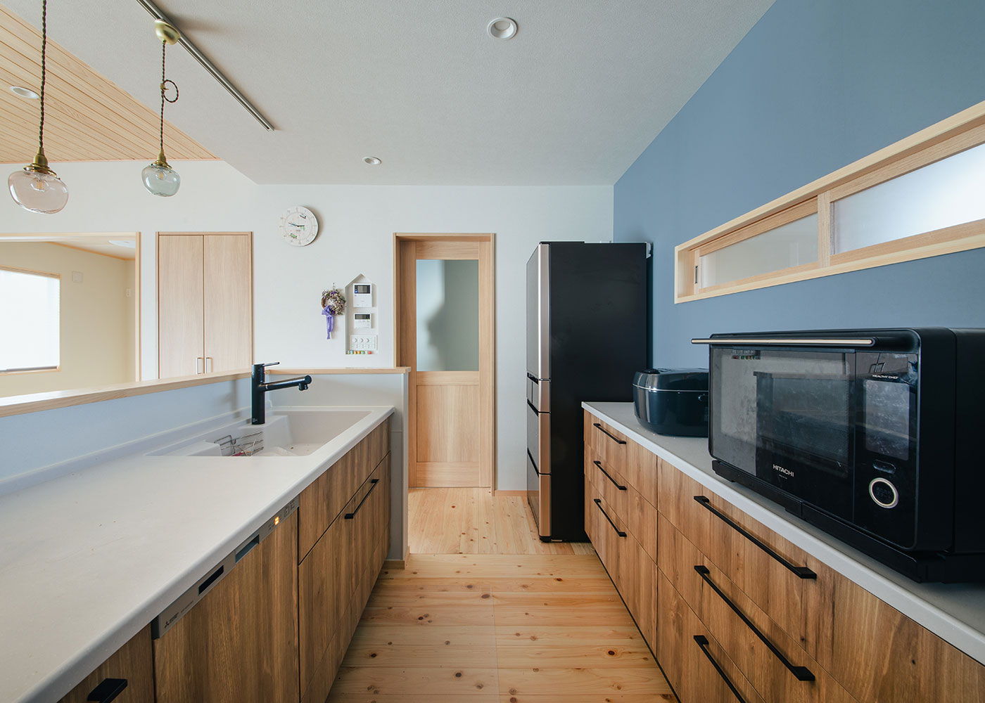 フジモクの家（富士木材）【子育て、間取り、平屋】ブルーグレーのクロスとブラウンのキッチンセットは奥さまのセレクト。右手の室内窓の向こうは洗面脱衣室になっており、さりげなく気配を伝える