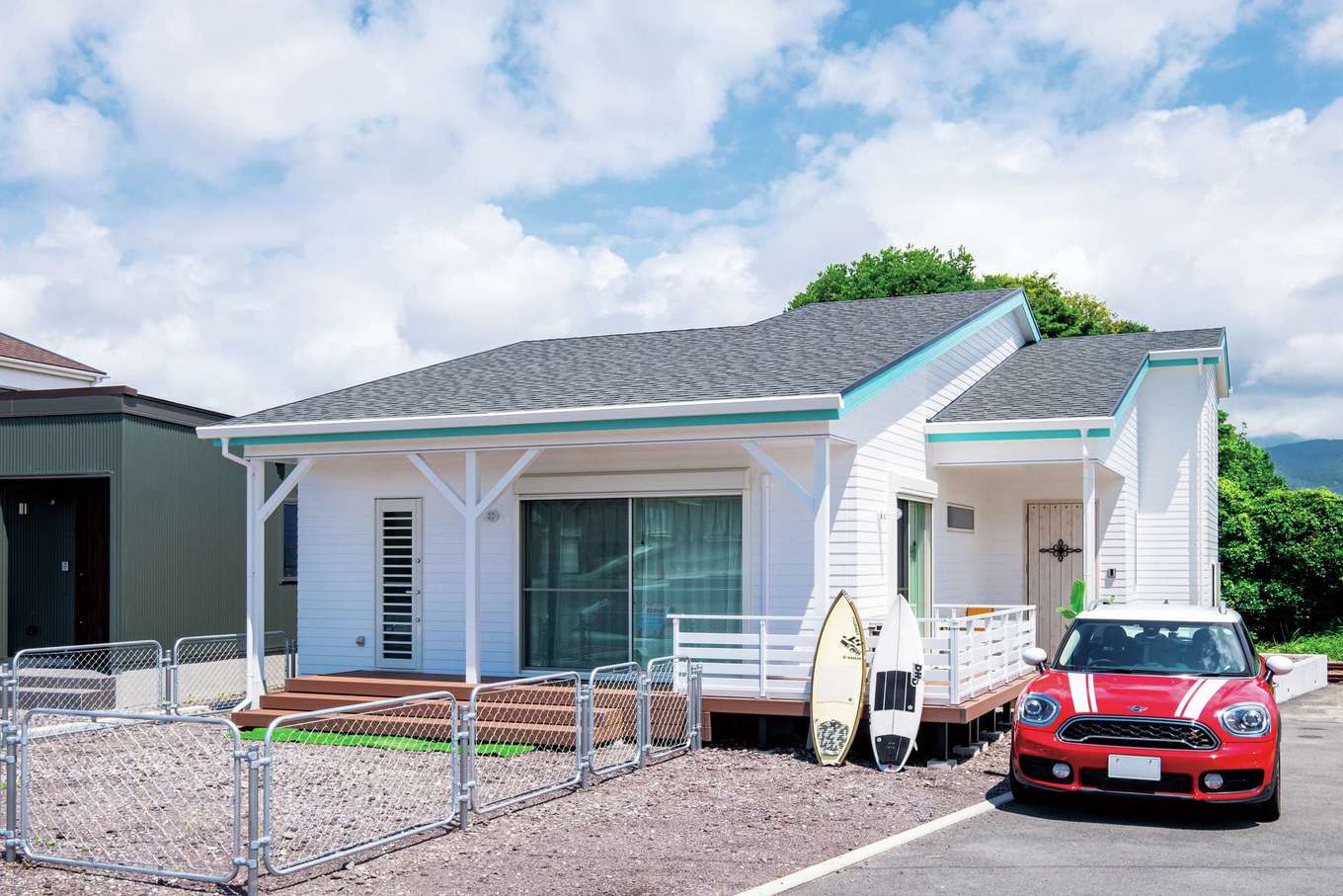 工藤建設【デザイン住宅、趣味、間取り】鎧張りの外壁は白の色味にこだわり、無塗装の建材に塗料を塗って“理想の白”に仕上げた。破風（はふ）のブルーが西海岸らしいさわやかなイメージを演出。駐車スペースも考慮しながらできるだけ庭を広くとった。日陰になるデッキは愛犬が休憩でき、BBQも楽しめる。愛犬が敷地外に出ないようデッキにもフェンスを付けた