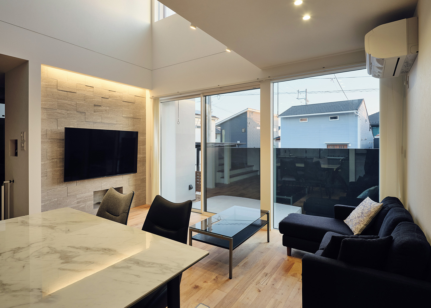 平屋建築ブルーハウス【デザイン住宅、間取り、ペット】リビングの開口部はF I X窓と引き違いの掃出窓を組み合わせ、ウッドデッキとの一体感を創出。アクセントウォールにはエコカラットを採用した。間接照明を灯すと、いっそうホテルライクな雰囲気に