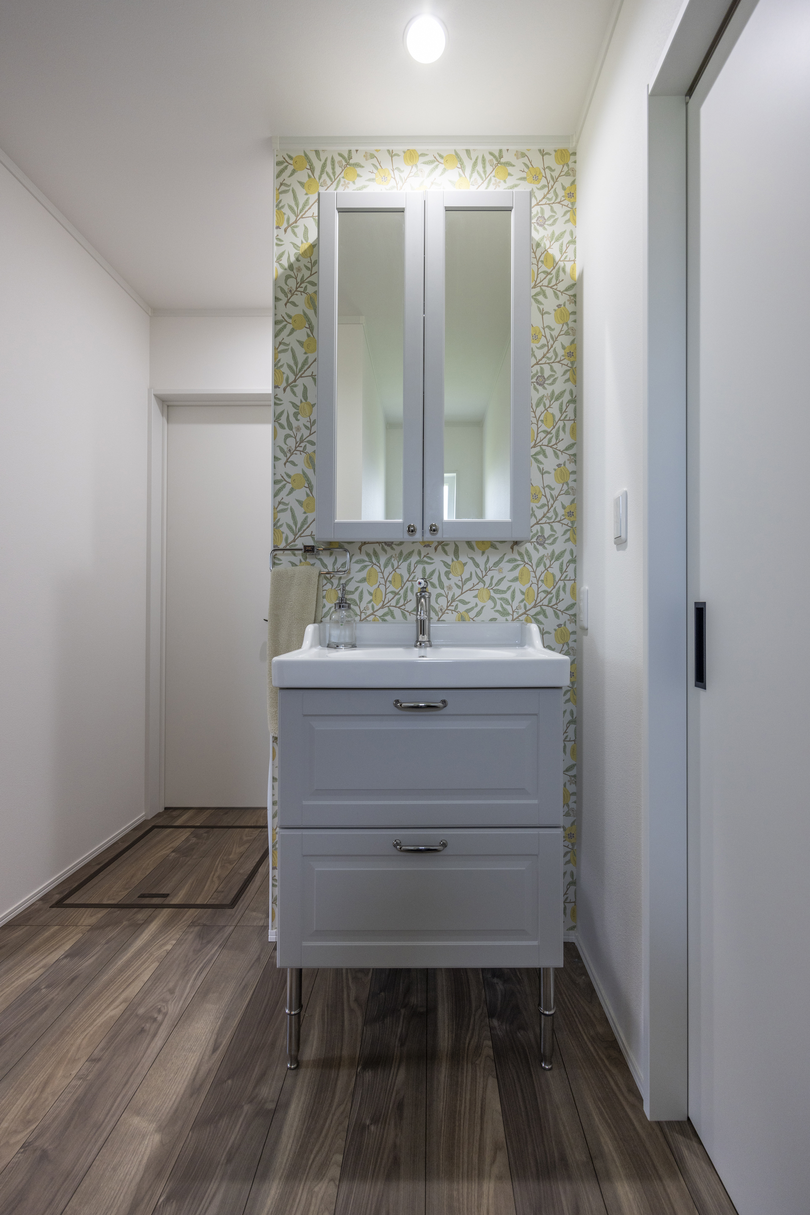 広島建設 セナリオハウス｜壁には温かみのある植物柄のアクセントクロスを使用した洗面スペース。シンプルで機能的なデザインが特徴で、使い勝手も良い。床の木目調が空間に優しさを加えています。