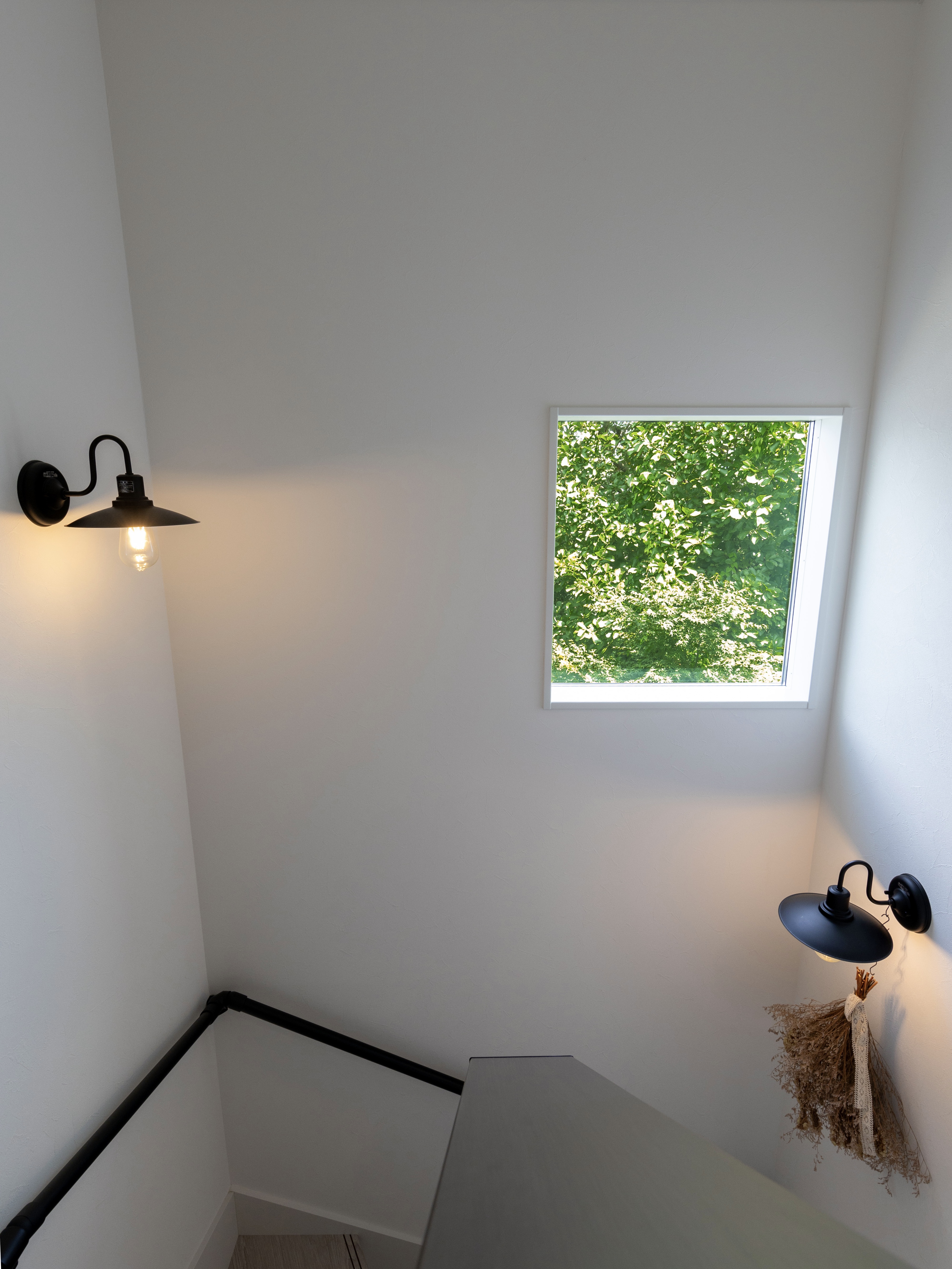 エスホーム｜白を基調とした明るい階段スペース。窓から自然光が入り込み、心地よい雰囲気を演出している。温かみのある照明がアクセント。