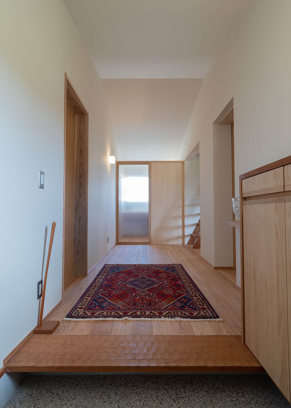 石牧建築【デザイン住宅、子育て、平屋】勾配天井の広々とした玄関ホールがお出迎え。上質なラグマットが明るい空間に彩りをプラスしている。框には同社気鋭の職人が名栗加工を施し、高級感を演出。つき当たりには洗面室があり、左には脱衣室と浴室、右に行くとパントリーとキッチンへつながるという合理的な間取り