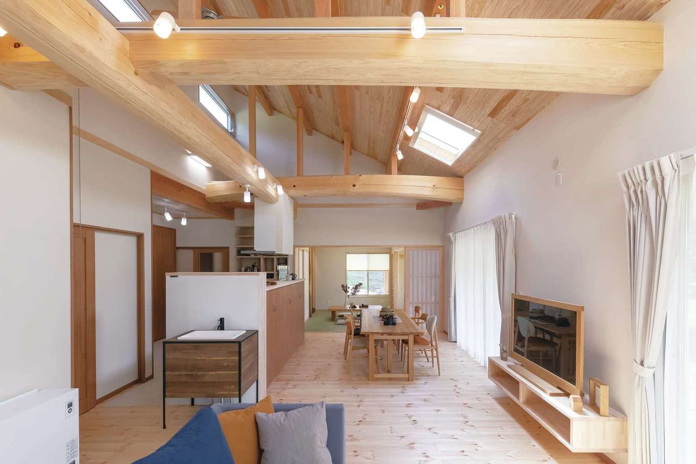 住まいるコーポレーション【デザイン住宅、自然素材、平屋】勾配天井に太い野物の梁がダイナミックに架かったLDKは、広さが約9畳。広い室内に明るさを十分確保するため、勾配天井にトップライトを2か所設置。リビングとダイニングの南面には全開口の掃出窓を設けてあり、常に美しい周囲の風景を見渡せる
