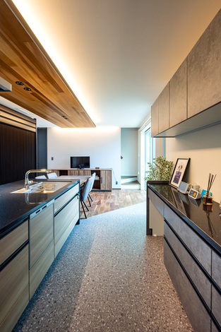 広々としたLDK空間。照明が部屋全体を柔らかく照らし、木目調の天井とフローリングが暖かさを演出している。モダンで洗練されたデザインのキッチンとリビングエリアが一体となり、家族の団らんに最適。