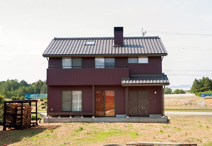 えんじ色の外壁と日本瓦を組み合わせ、軒天やデッキのブラウンを差し色にした個性的な外観。庭先にはご主人と『戸田工務店』のスタッフ達で造った薪棚もある