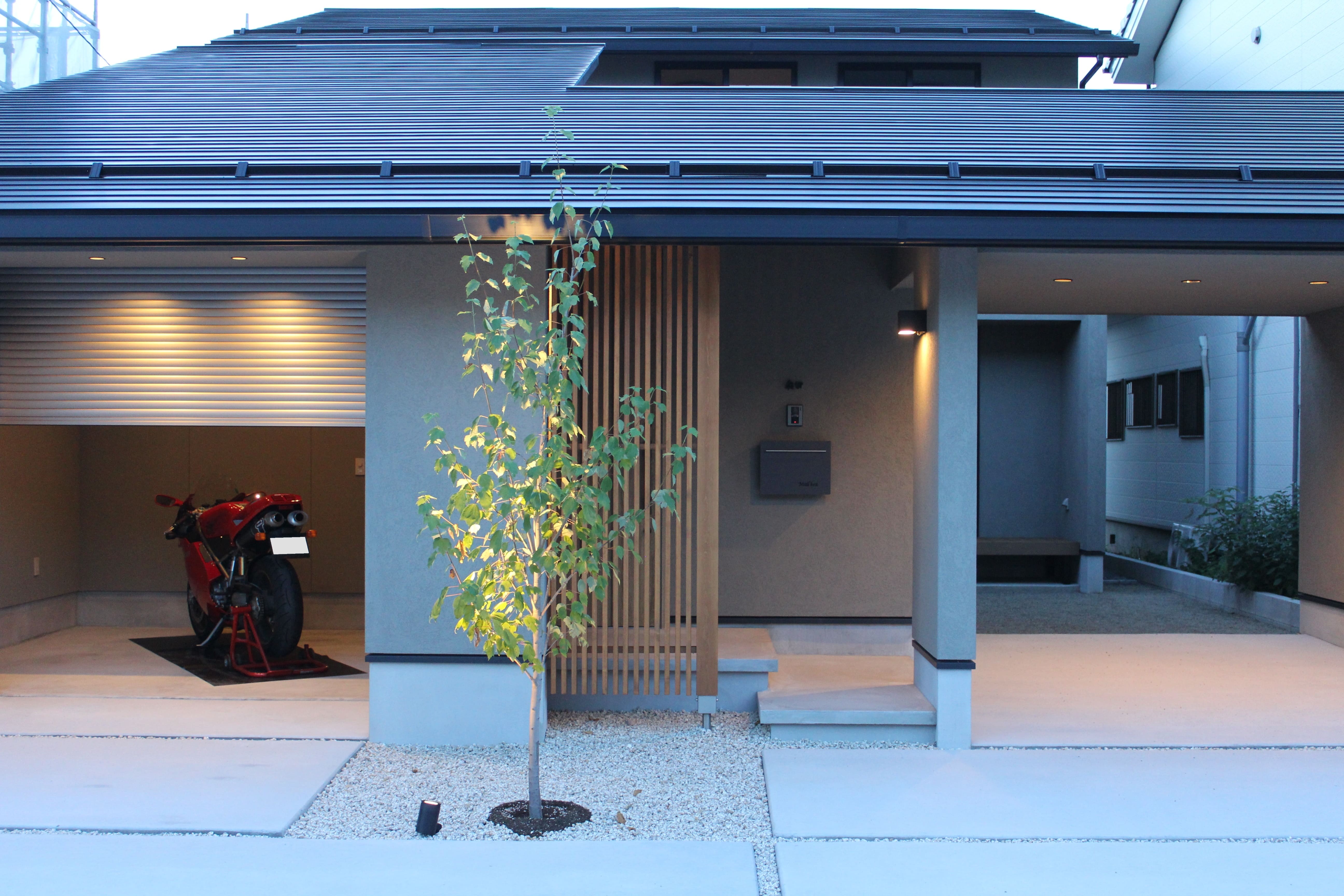 安田住宅｜安田建築設計室｜現代的なスタイルの家の外観。ガレージにバイクが停められ、正面にはシンプルで洗練されたデザインのエントランスが見える。舗装されたアプローチには小さな植栽が映える。