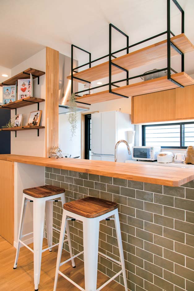 仲田工務店【デザイン住宅、子育て、間取り】タイル壁に木の天板のカウンター、木とアイアンの吊り棚、家族写真を飾る棚など、奥さまの要望を形にしたキッチン。ぐるぐる回れるレイアウトも気に入っているそう