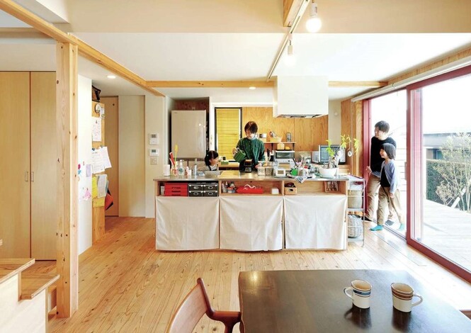入政建築｜スギの床の柔らかな感触と構造用合板の飾り気のない雰囲気が心地よいLDK。大きな窓を介して緩やかにウッドデッキとつながっているので、いつでもすぐに「デッキでご飯」ができるところも夫婦のお気に入り。造作の木のアイランドキッチンは、既製品を入れるか迷った末に取り入れたこだわりのもの。コストダウンのためにあえて収納扉を付けず、布で隠すというアイデアも秀逸。「暮らしを育てる」ことを楽しむ夫妻にぴったりの空間となっている