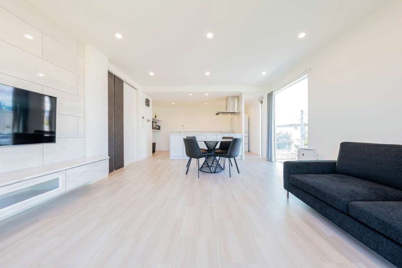 大成工務店【デザイン住宅、収納力、高級住宅】ダイニングの大きな窓が開放感を生むLDK。住居スペースのインテリアも白を基調にした。家具をモノトーンで統一し、すっきりとした印象に。間接照明やテレビ側壁面のパネルは、白一色の中に絶妙のニュアンスをプラスしている