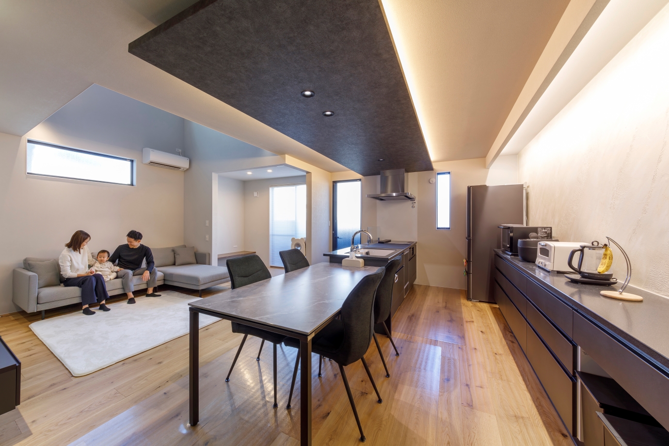 nattoku住宅【デザイン住宅、収納力、間取り】スタイリッシュ＆モダンな雰囲気にコーディネートされた20畳のLDK。キッチン背面の壁にイタリア漆喰「ユーロスタッコ」を施しアートな雰囲気を演出。下がり天井、間接照明との相性もぴったり。グレーのダイニングテーブルセット、L字型のソファは『nattoku住宅』のオリジナルアイテム