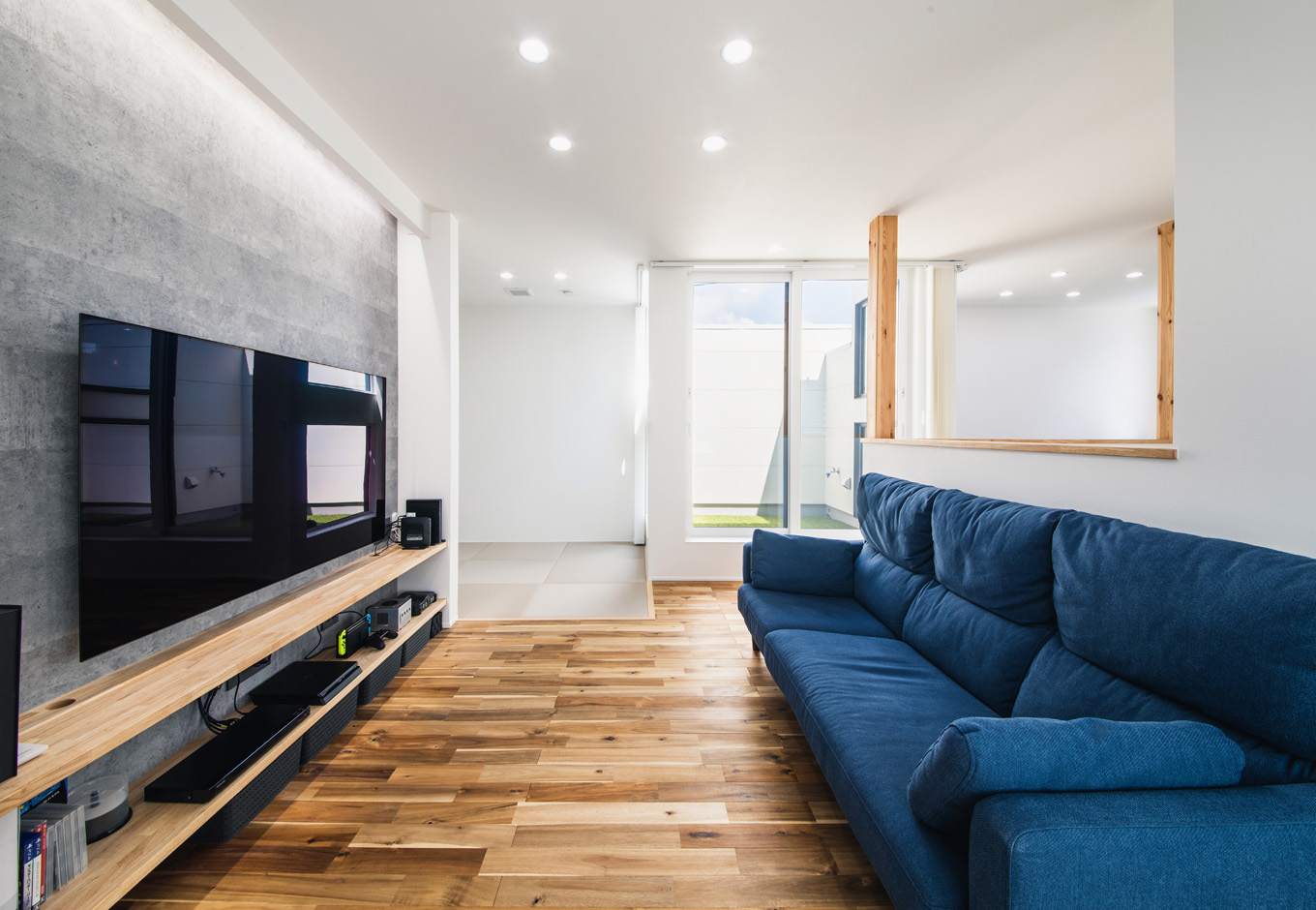 SEIEN ARCHITECT【趣味、建築家、1000万円台】夫妻がくつろぐリビングはゆとりの広さ。造作のT Vボードを壁一面に設け、大型テレビの前にベッド代わりにもなりそうな3人掛けのソファを置いてある。無垢のアカシアの床が素足に心地よい