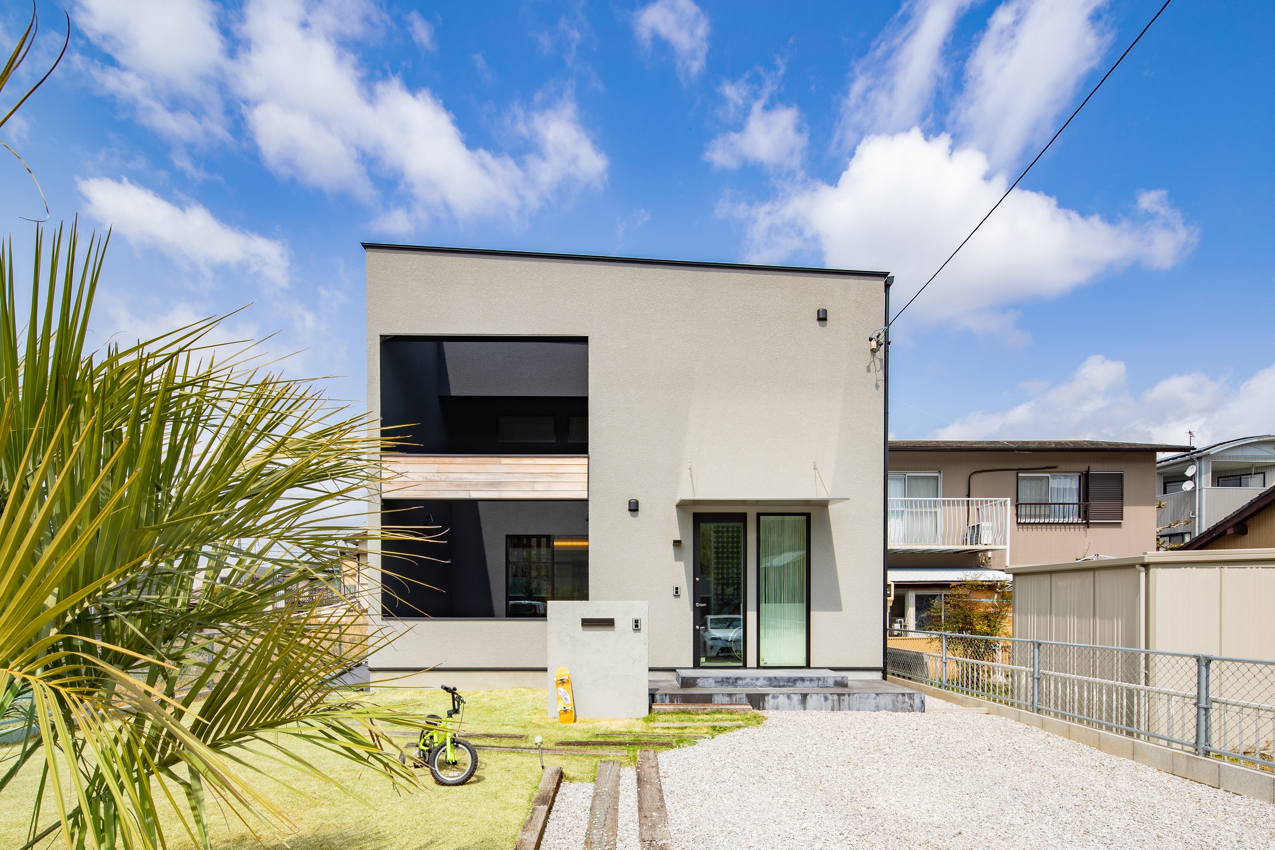 S.CONNECT（エスコネクト）【デザイン住宅、子育て、建築家】ヤシの木と青空が似合うスタイリッシュな外観。グレーの外壁に木目のアクセントが引き立っている。デッキを隠すように外壁を設けたことで、内と外の中間のような丁度いお籠もり感を叶えた