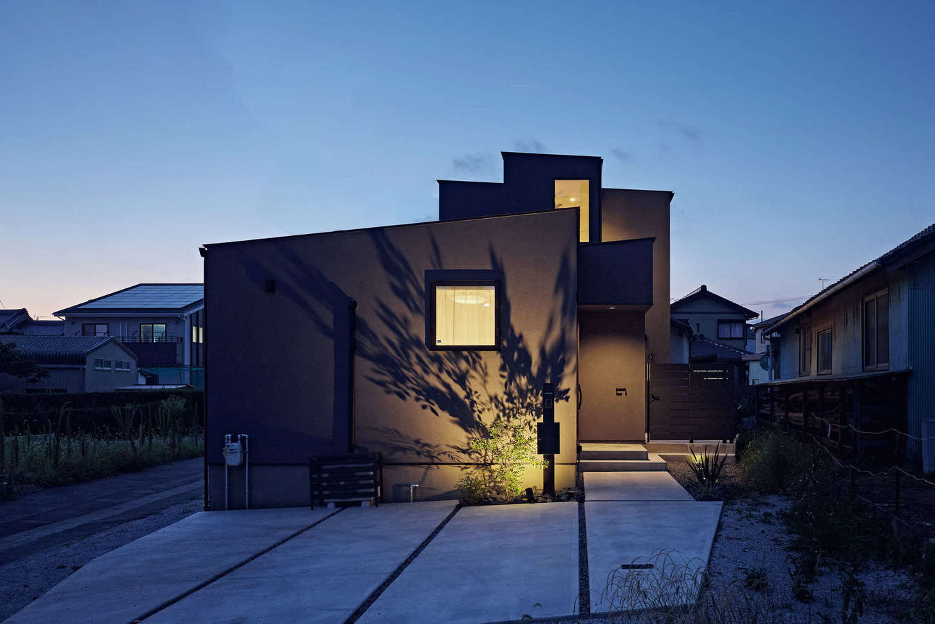 平屋建築ブルーハウス【デザイン住宅、建築家、平屋】濃いグレーの塗り壁とスタイリッシュなフォルムが夜の照明に映える外観。塗り壁は、「高本コーポレーション」の人気カラーをチョイスした。正面の正方形窓の先は寝室のため、高い位置に配して周囲の視線をカット