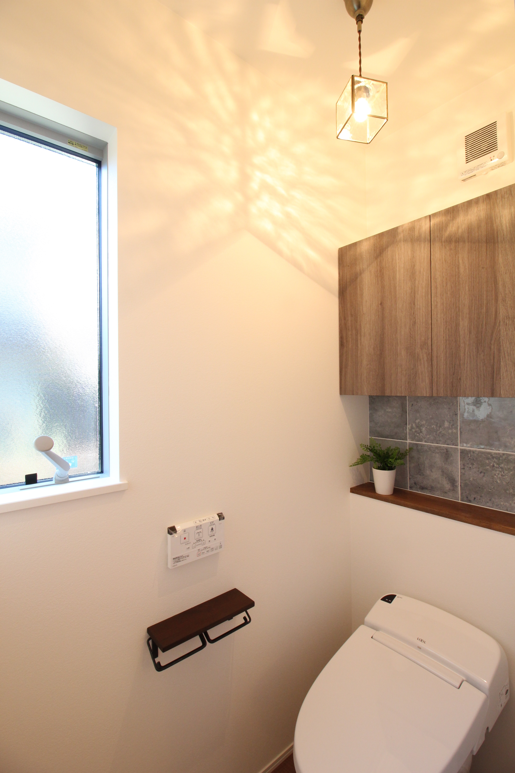 小野田産業～designers house project～｜温かみのある照明と木目調キャビネットが特徴のトイレ空間。窓から差し込む自然光が明るさを提供し、植物がアクセントを加えている。