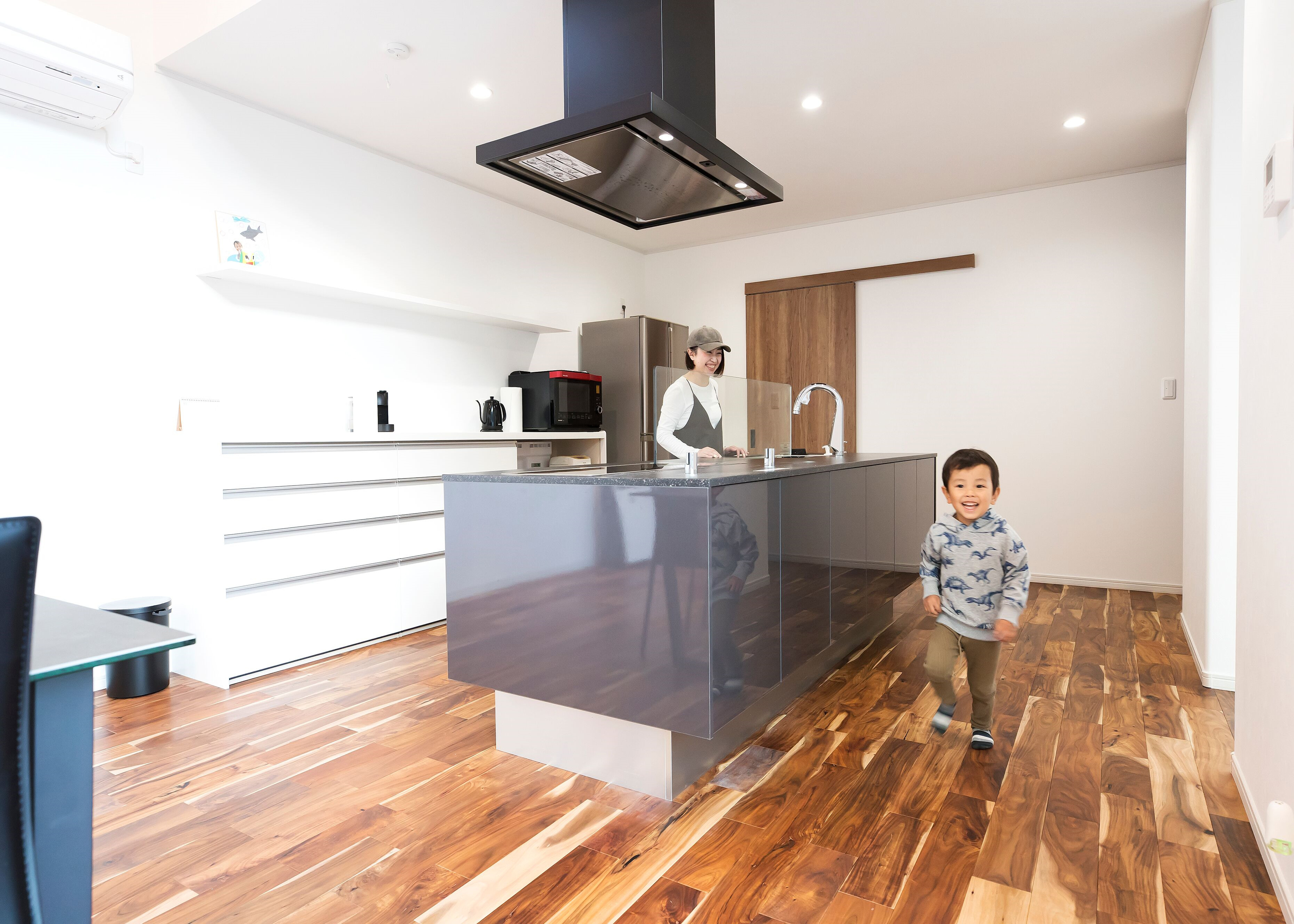 子育て安心住宅＆デザインラボ【デザイン住宅、間取り、平屋】アイランドキッチンはワークスペースを広く取り、家族一緒に料理作りを楽しめるようにした。背面の収納は壁と同じ白にして明るくスッキリとした印象に。収納は取り出しやすさを重視して、引き出しタイプを選択。キッチンの奥にはパントリーがある
