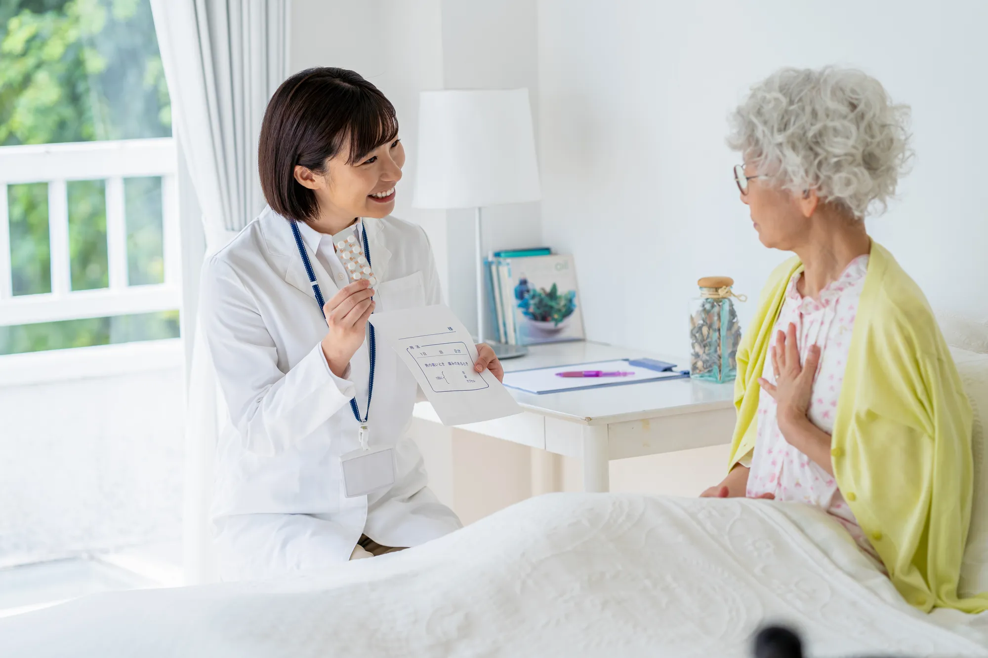 白衣を着た女性が病室で薬袋を白髪の女性に見せている