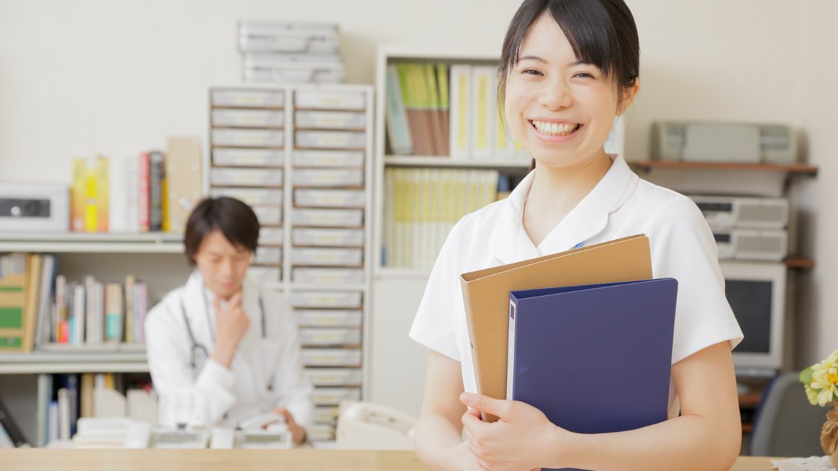 白衣を着た女性が笑顔で版を持って立っている