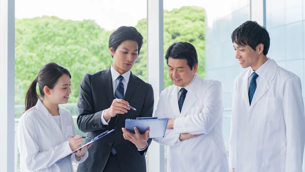 白衣を着た男性と女性がスーツの男性を囲って説明を聞いている
