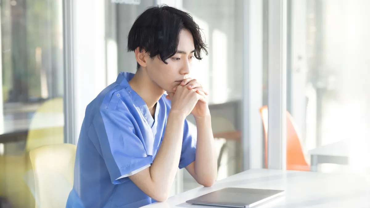 白衣を着た男性がパソコンの前に座って悩んでる