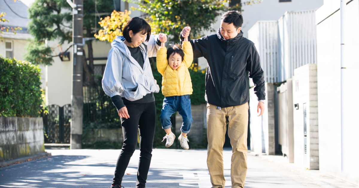 親が子どもの両手を持ち、住宅街の道路で子どもを持ち上げながら歩いている。