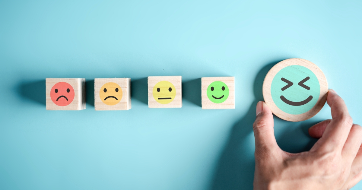 水色の背景の上に、悲しい顔→不満顔→無表情→笑顔の絵が描かれた木製キューブが並び、右側で手が大きな満面の笑顔の木製アイコンを持っている