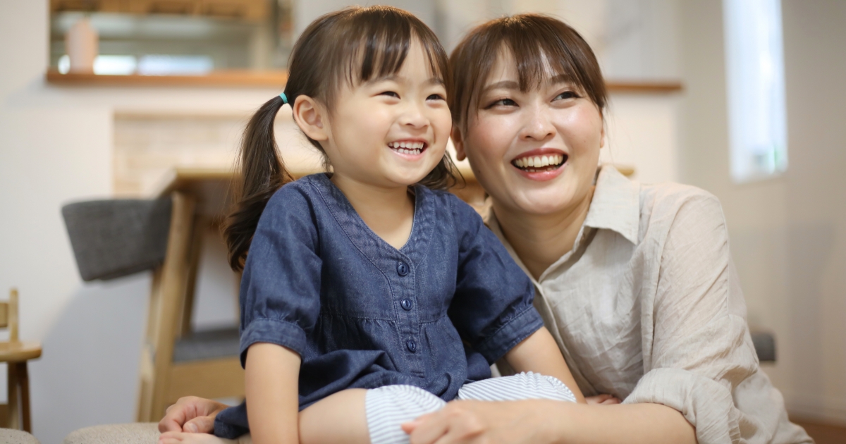笑顔の母親と幼い女の子が寄り添って座り、室内で楽しそうに同じ方向を見ている様子