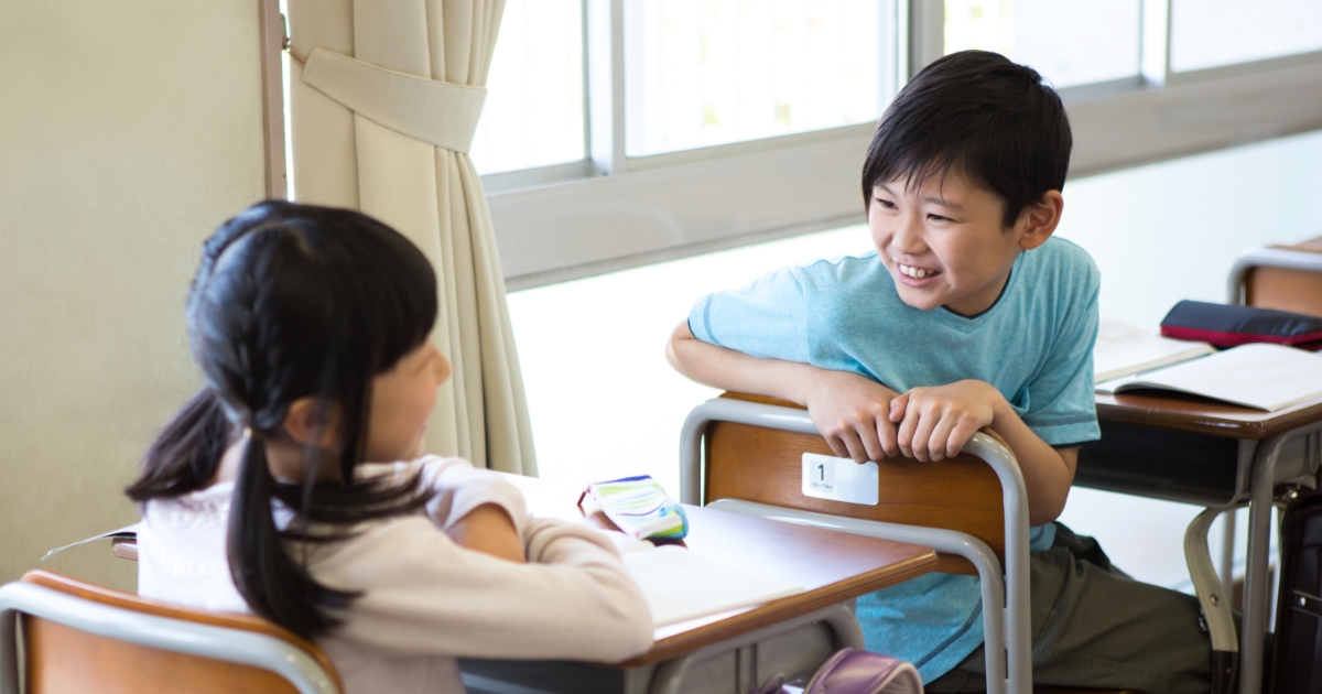教室で席を向かい合わせた小学生の男女が、笑顔で会話している場面