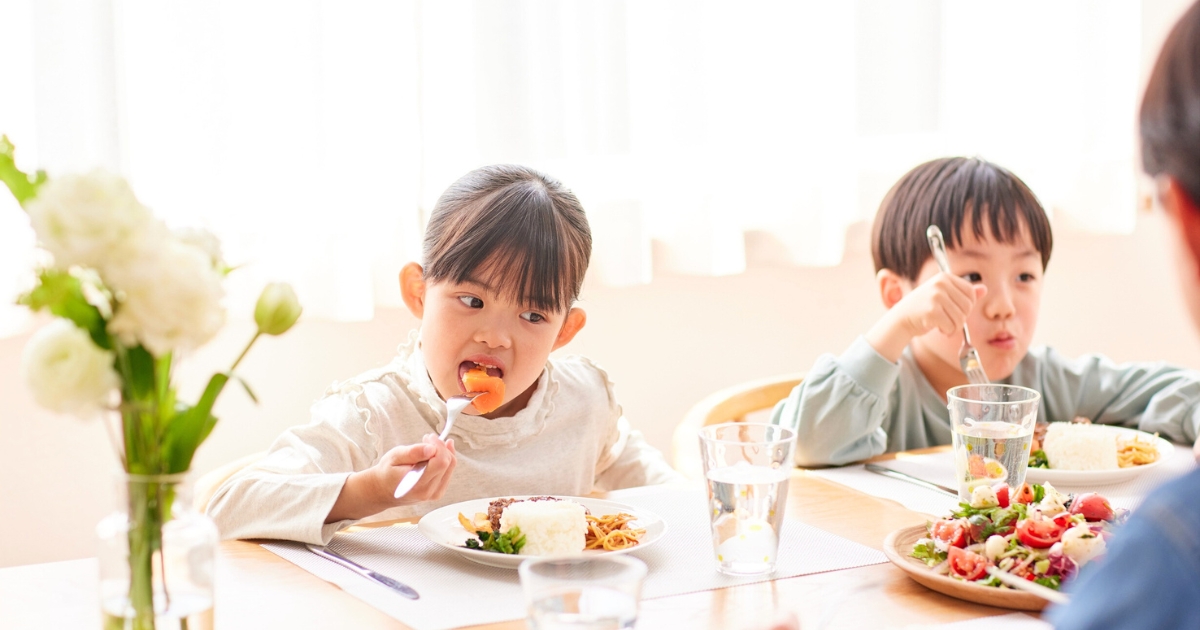 明るい食卓で、幼い女の子と男の子がフォークを使って食事をしている。