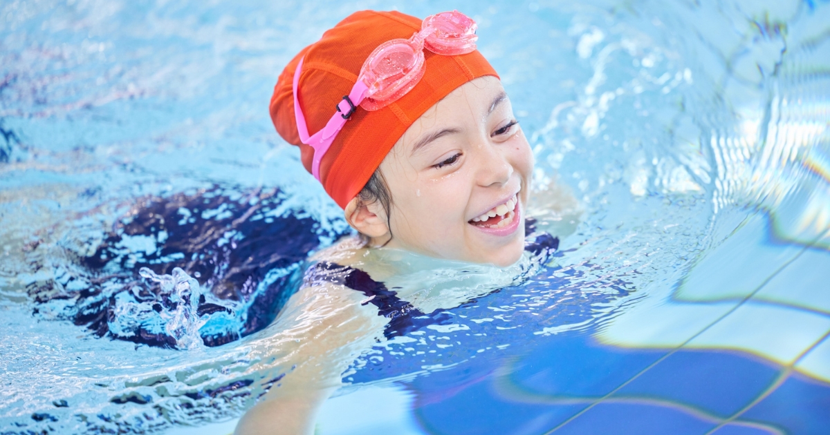 オレンジ色のスイムキャップとゴーグルを着けた子どもが、プールで笑顔で泳いでいる様子