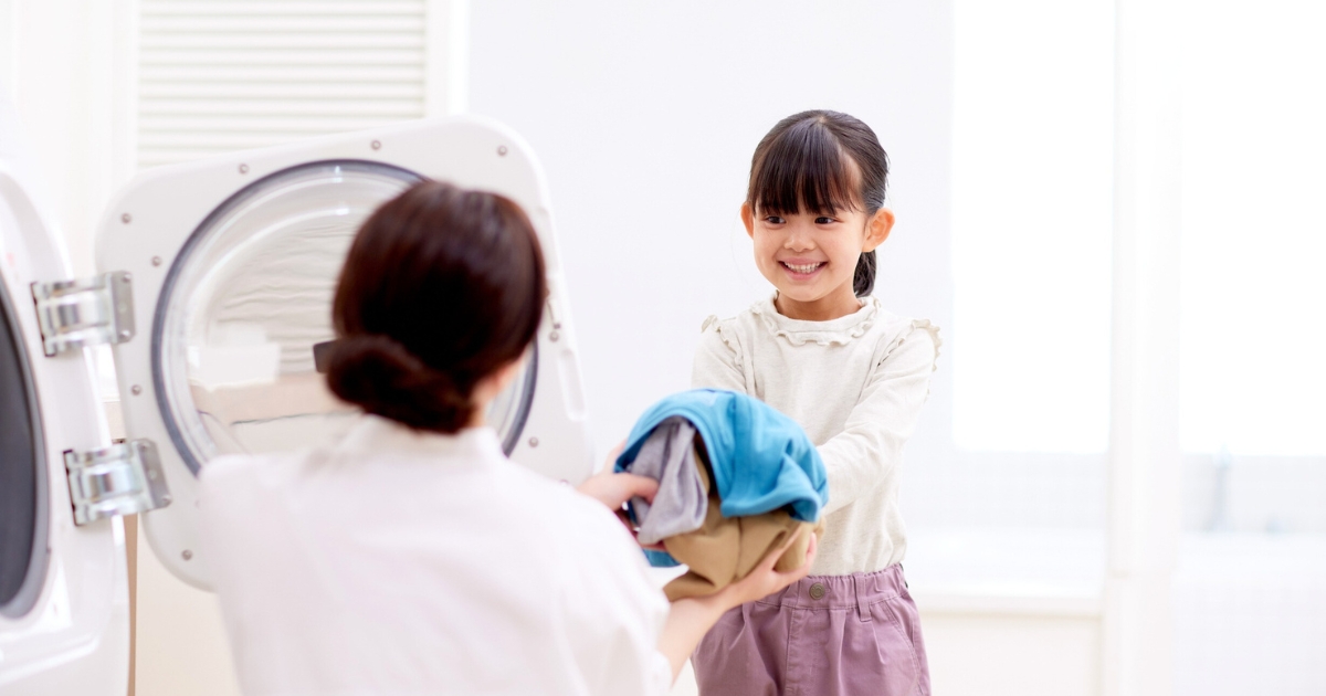 洗濯物を手にした女の子が、洗濯機の前で待つ大人に笑顔で衣類を渡してお手伝いをしている様子