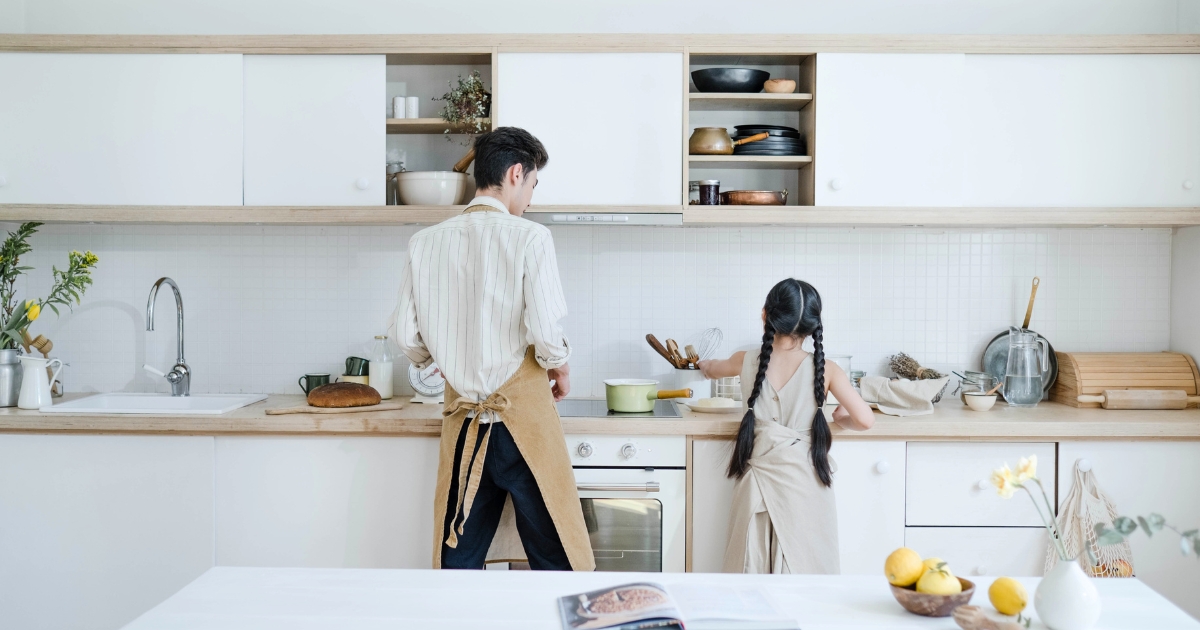 明るく整ったキッチンで、エプロンを着けた父親と三つ編みの子どもが並んで立ち、調理台に向かっていっしょに料理をしている後ろ姿