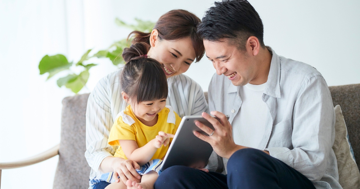 ソファに座り、父親が持つタブレット画面を指さして楽しそうに見つめる親子3人
