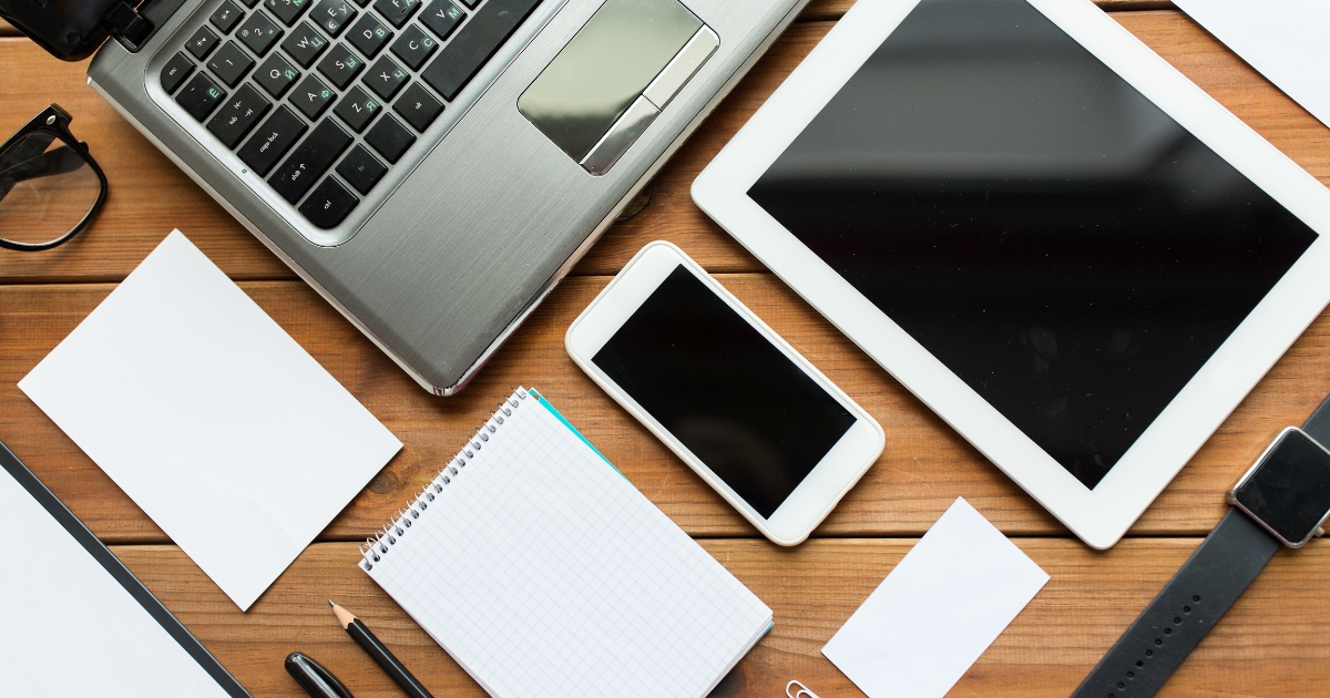 木の机の上にノートパソコン、タブレット、スマートフォン、メモ帳などのデジタル機器と文具が並んでいる様子