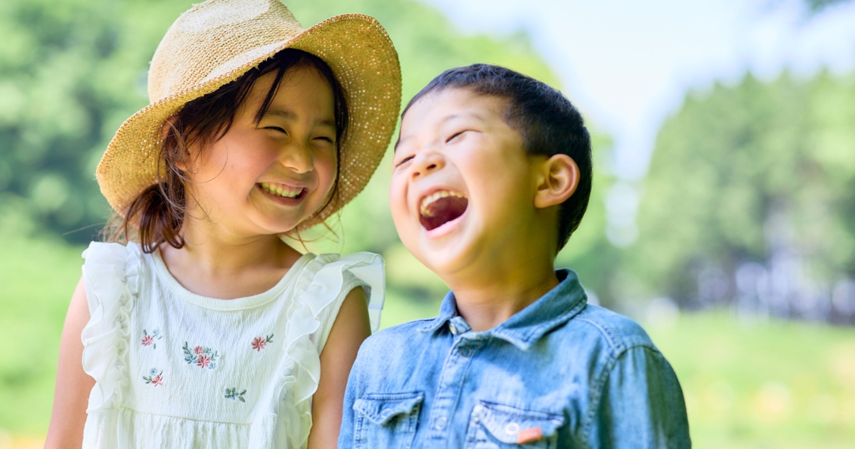公園で帽子をかぶった女の子と男の子が顔を見合わせて楽しそうに笑っている様子。子どもの自然体験・友達との関わりのイメージ