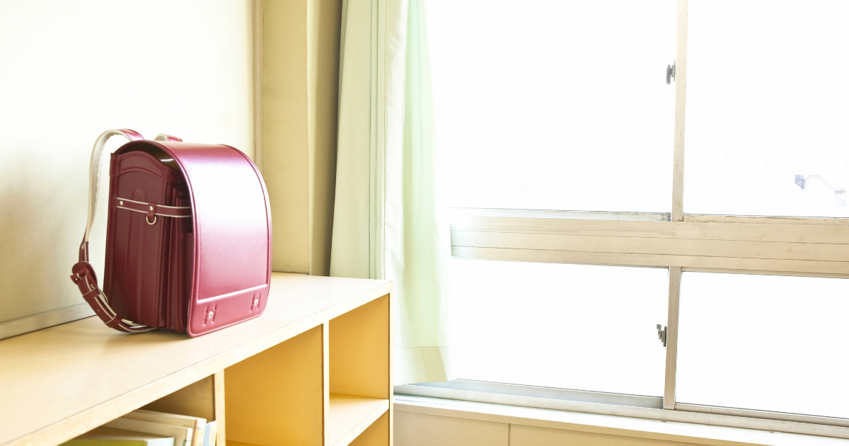 明るい光が差し込む教室の窓際で、木の棚の上に置かれた赤いランドセル