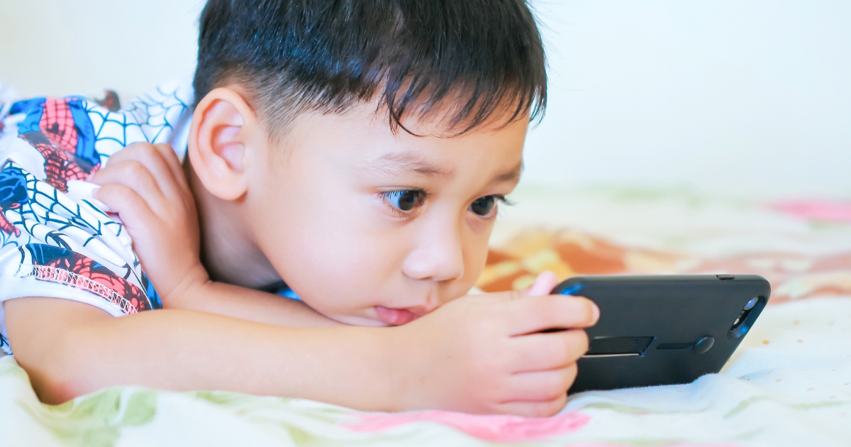 ベッドにうつ伏せになりスマートフォンを操作する小さな男の子。画面に集中している様子
