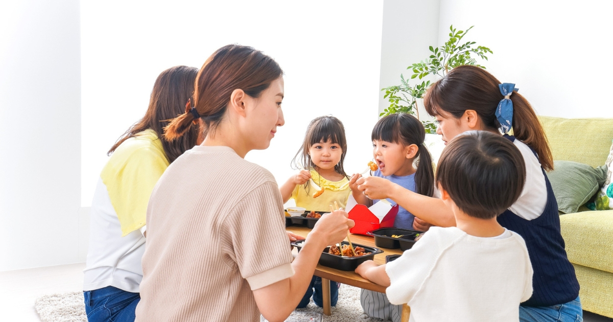 リビングのローテーブルを囲み、複数の親子が一緒に食事を楽しんでいる様子。子どもたちに大人が食べ物を取り分け、明るい室内で団らんの時間を過ごしている