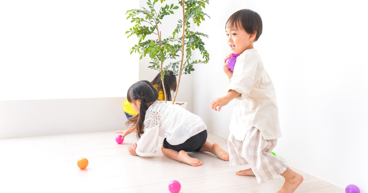 室内の白い床の上で、カラフルなボールを拾ったり投げたりして元気に遊ぶ3人の子どもたち