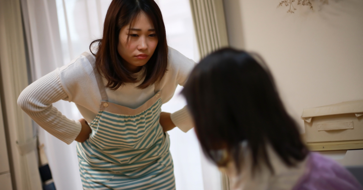 心配そうな表情の女性が、前かがみになって座る子どもに話しかけている室内の様子。
