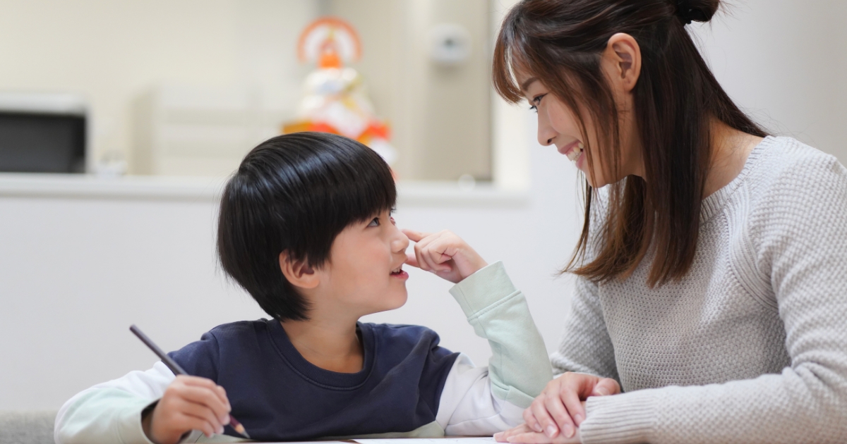 笑顔の母親と向かい合いながら、机で勉強する男の子。