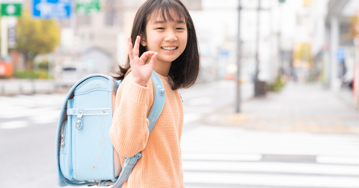 水色のランドセルを背負った女の子が、街中の横断歩道付近で笑顔を見せながらOKサインをしている様子