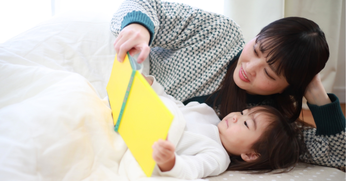 布団の中で横になりながら、黄色い絵本を一緒に読む母親と小さな子ども