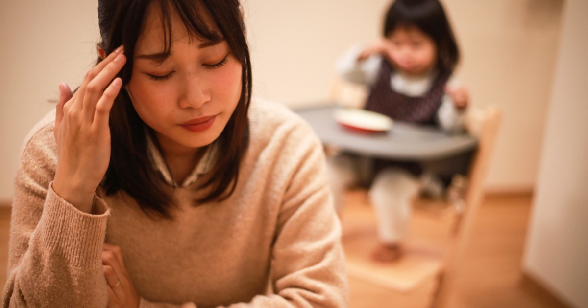目を閉じてこめかみに手を当て、育児に疲れ切った様子の母親と、奥のベビーチェアに座る子ども