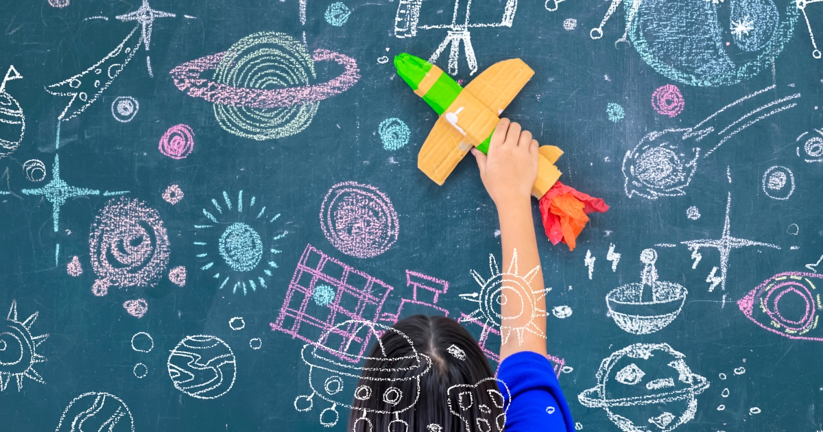 宇宙の絵が描かれた黒板の前で、手づくりのロケットを掲げる子ども