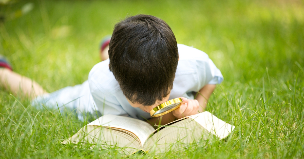 芝生の上に寝転がり、虫眼鏡で本を読む男の子