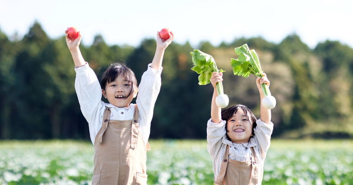 畑で収穫したトマトとカブを両手で高く掲げて、笑顔を見せる2人の女の子