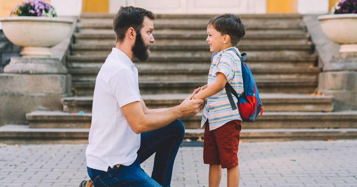 階段の前で、男性がしゃがんで子どもと向き合い、手をつないでいる様子。子どもはリュックを背負い、笑顔で男性を見ている。